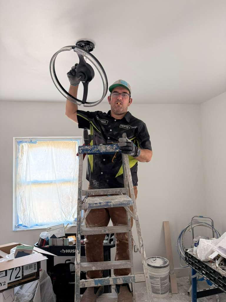 An electrician on a ladder installing a ceiling fan or light fixture. The room appears to be under renovation with a window covered in plastic and various tools and materials scattered around. The electrician is wearing gloves and looking towards the camera.