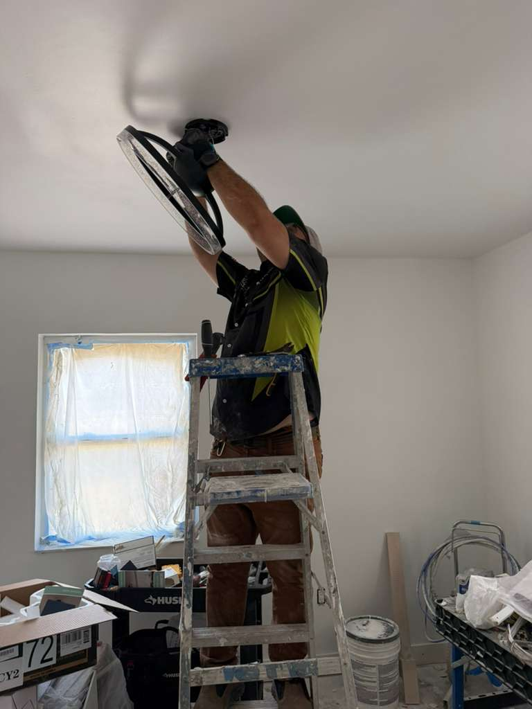 A worker on a ladder installing a new light fixture on the ceiling during a remodeling project. The room appears to be freshly painted with white walls. There are boxes and tools on the floor near the ladder, suggesting ongoing work.