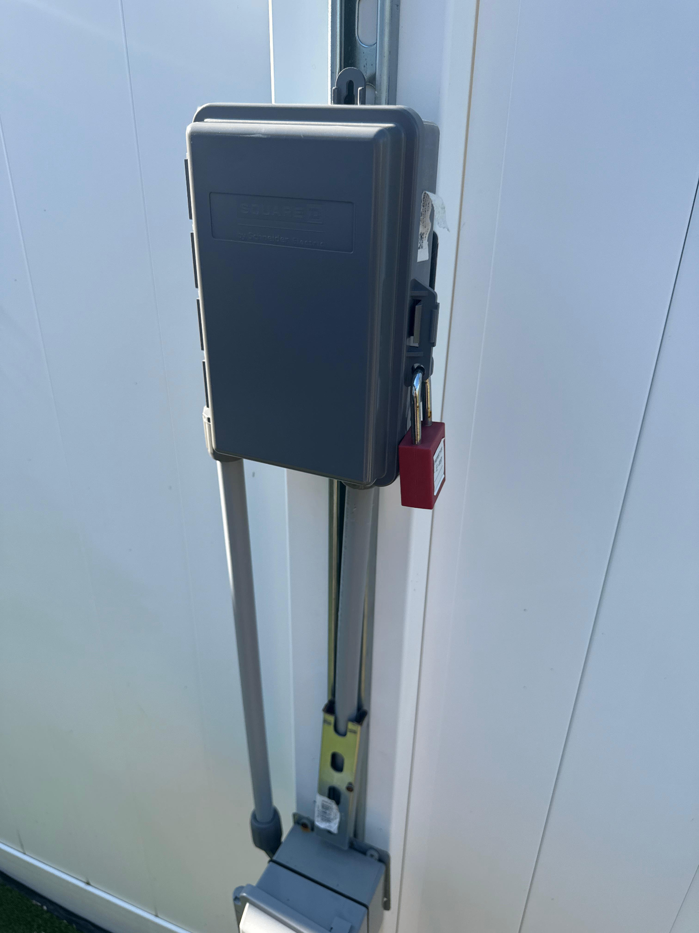 An outdoor electrical box, made by Square D by Schneider Electric, is securely locked with a red padlock. Gray conduits run from the box to an unknown destination, likely underground. The box is mounted on a white, possibly exterior, wall.