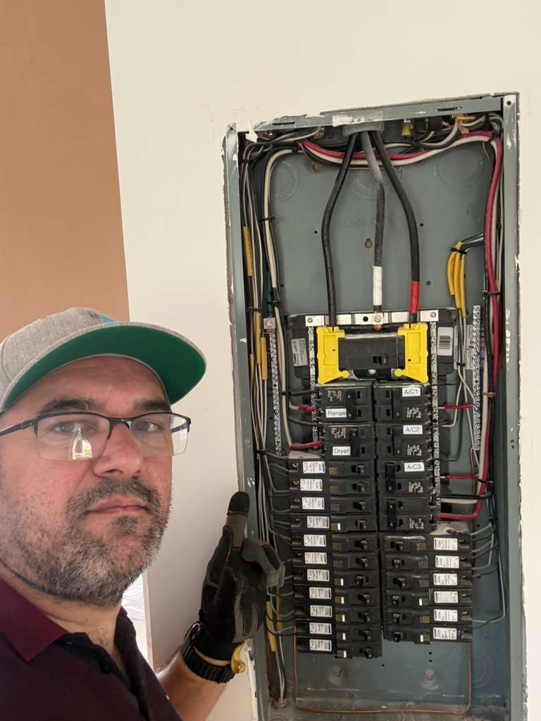 A man stands next to an open electrical panel with many circuit breakers, some labeled (e.g., Range, Dryer, A/C). The wiring is visible and appears to be newly installed or upgraded. The image shows a close-up of the electrical work.