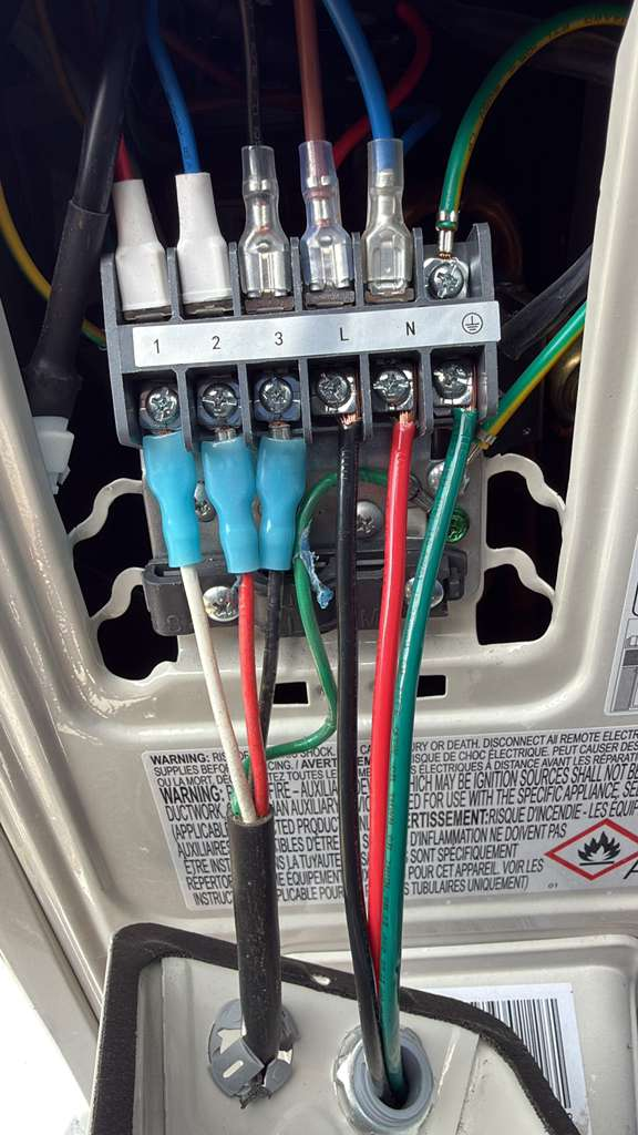 Wiring for an AC/Mini-Split unit showing multiple colored wires connected to a terminal block. The wires are secured with connectors, and the terminal block is labeled with numbers, L, and N, and a ground symbol. Warning labels are visible in the background.
