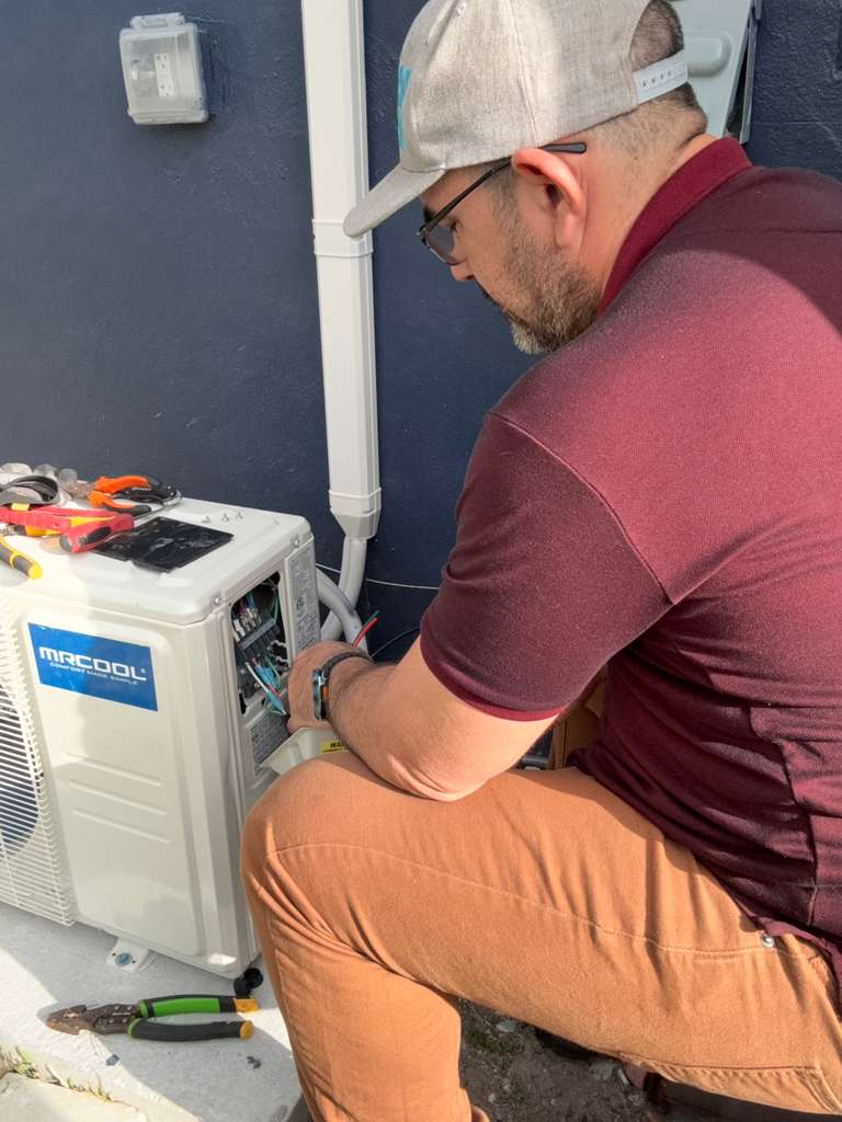 A man is installing an AC / Mini-Split unit. The unit is white and has "MRCOOL" written on it. The man is wearing a maroon shirt, brown pants, and a grey hat. He is working on the electrical wiring of the unit. There are tools next to him.