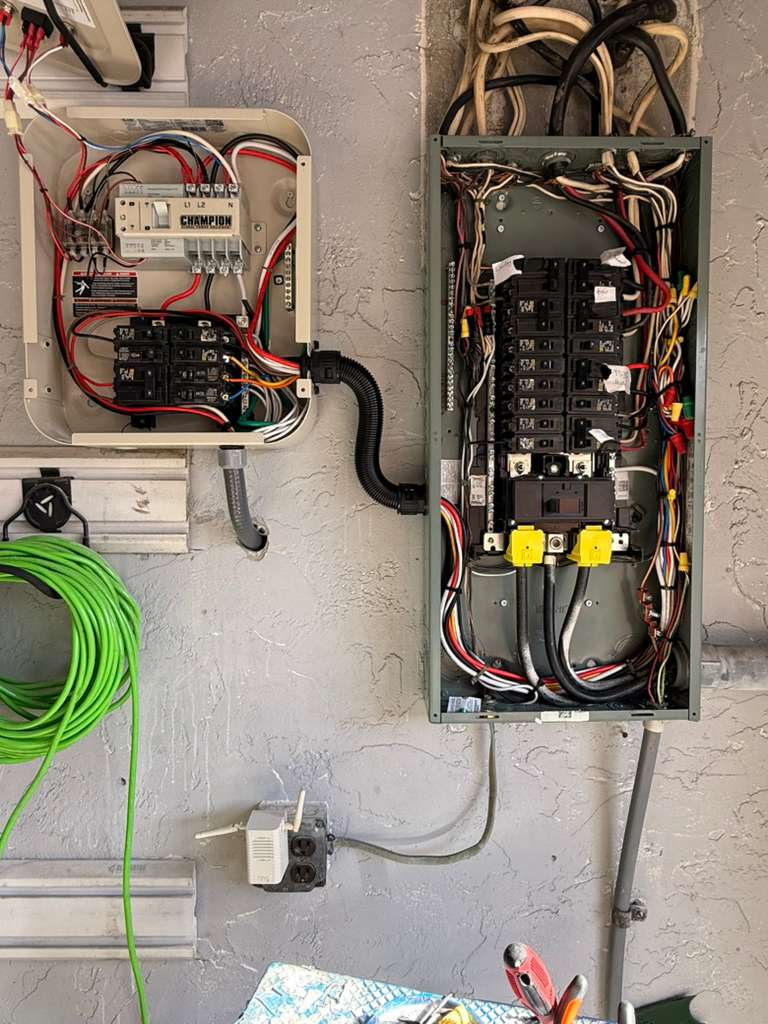 This image shows an electrical panel upgrade with a new transfer switch (Champion brand) being installed next to an existing electrical panel. The wiring is visible and appears to be a professional installation.