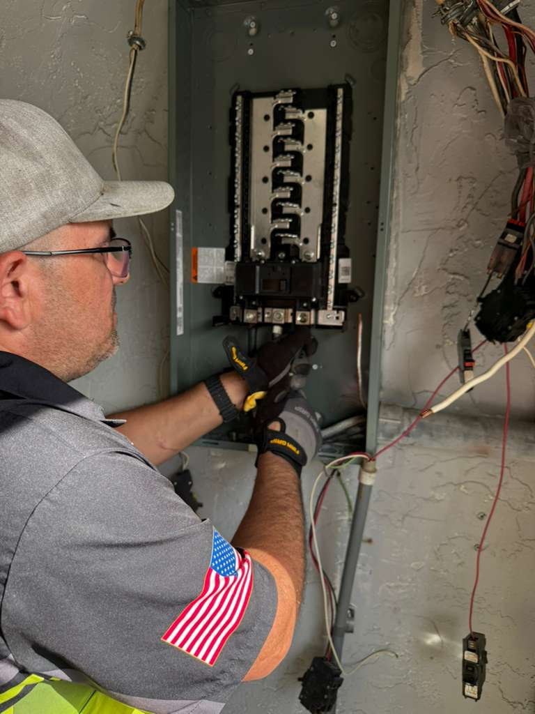A close-up shot of an electrician working on a residential electrical panel. The panel is open, revealing the breakers and wiring. The electrician is wearing gloves and appears to be connecting or disconnecting wires.