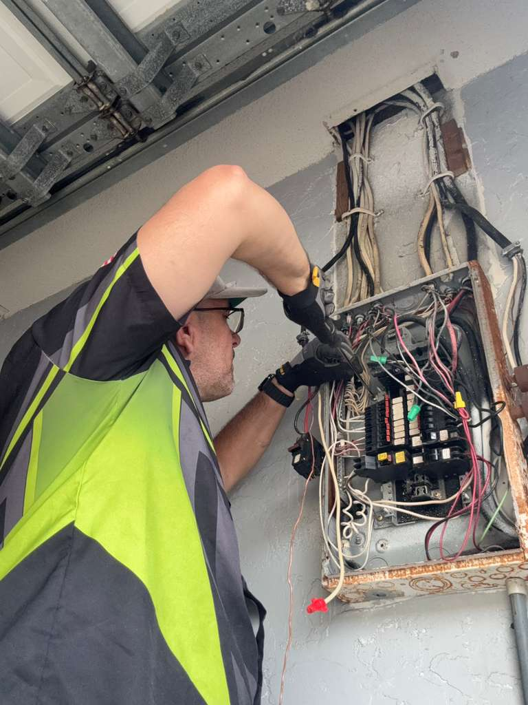 A worker upgrading an electrical panel, with numerous wires and circuit breakers visible. The panel itself appears somewhat old and possibly corroded, suggesting a need for the upgrade.