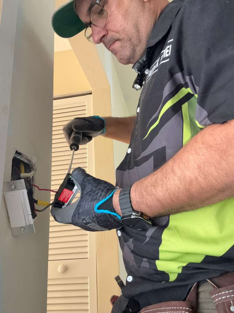 An electrician is installing a GFCI outlet, connecting wires with a screwdriver. The outlet is partially installed in the wall, with red, yellow, and black wires visible.