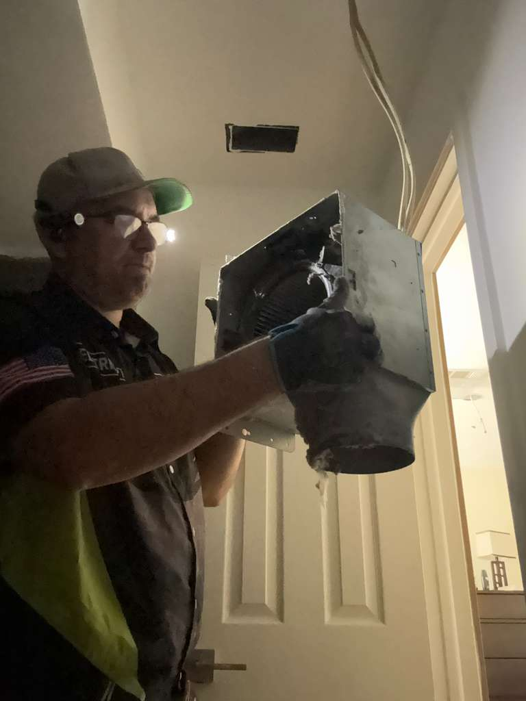 Contractor holding an AC / mini-split unit, with wiring visible and a hole in the ceiling where it will be installed. The contractor is wearing a headlamp and safety glasses.