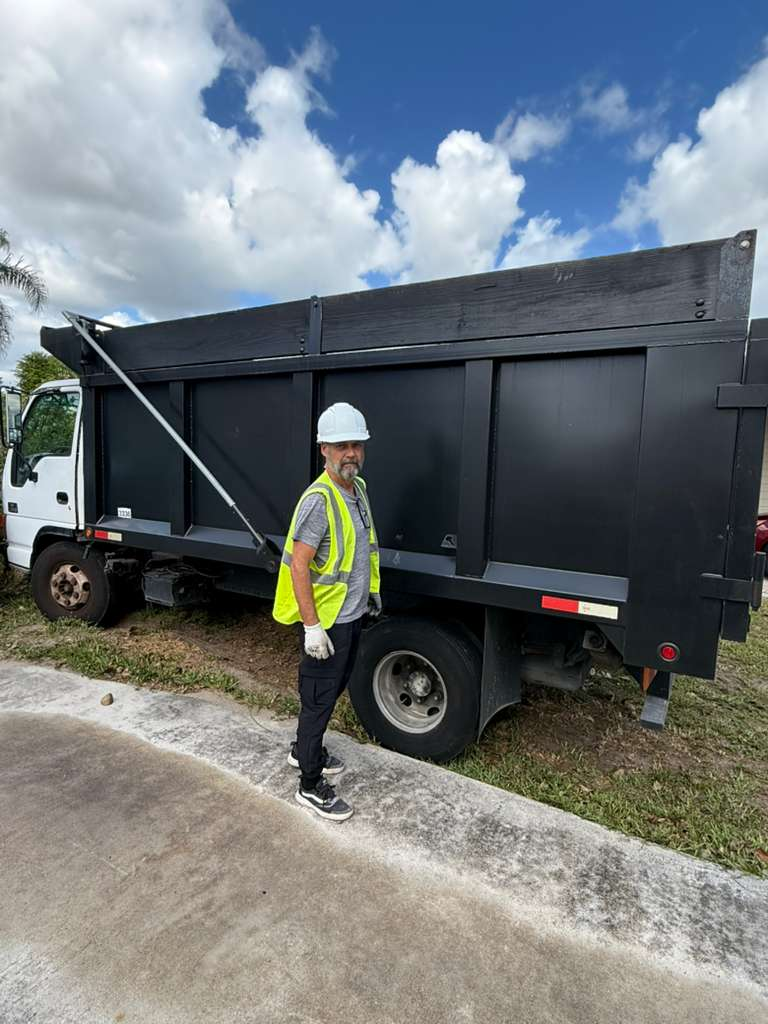 A man in a hard hat and safety vest standing next to a large dump truck, likely for debris removal.