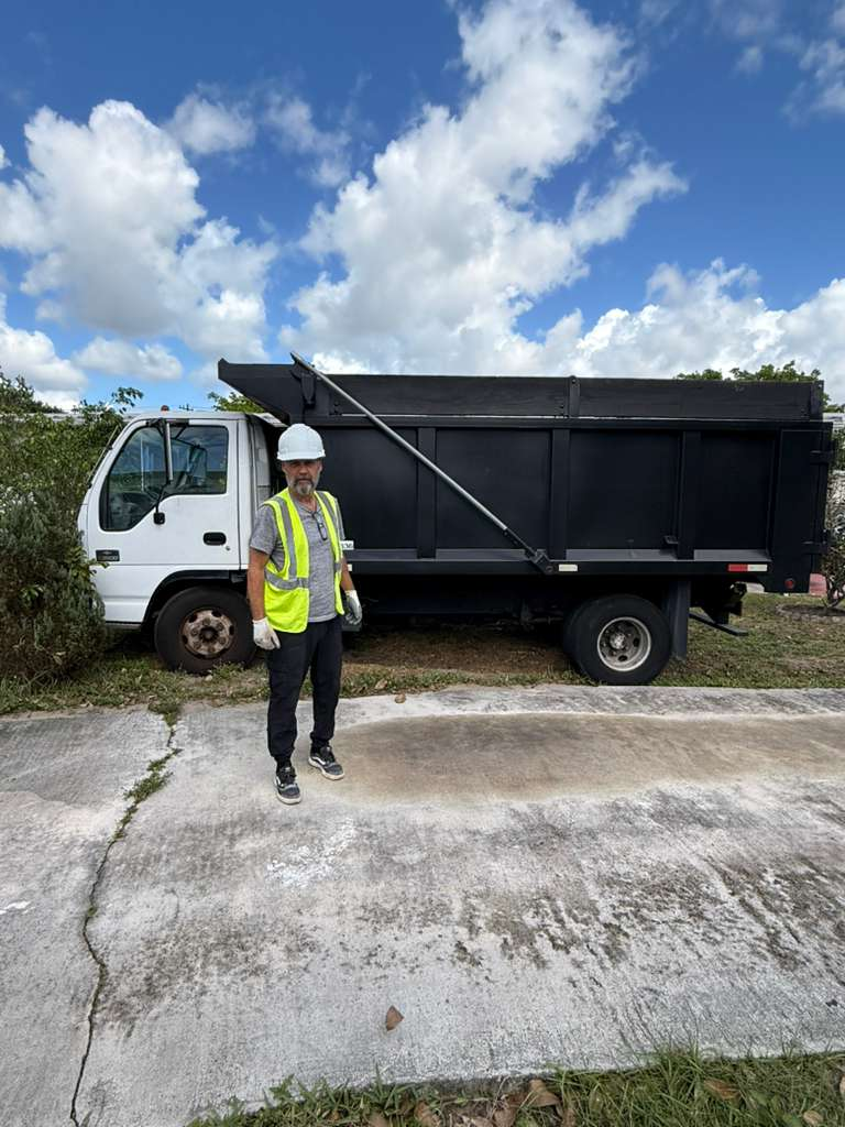 A man in a hard hat and safety vest standing next to a dump truck, which is likely used for debris removal.