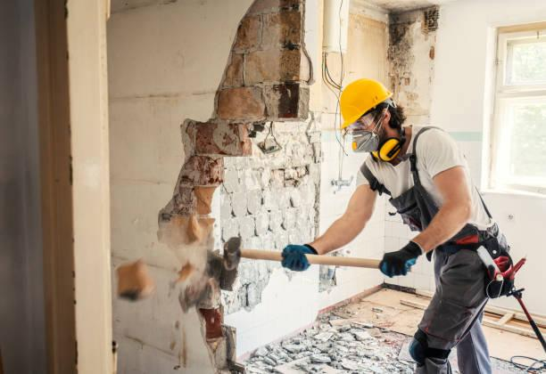 A worker wearing a hard hat, respirator, and ear protection uses a sledgehammer to demolish an interior wall, creating a large opening and scattering debris on the floor.