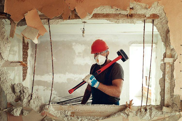 Worker with sledgehammer breaking through a wall, revealing rebar and debris. Plaster and wallpaper are peeling from the ceiling.