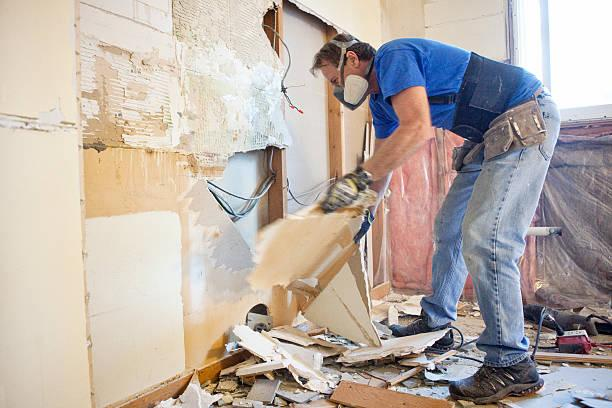A worker is demolishing an interior wall, creating debris. The wall appears to be made of lath and plaster or drywall, revealing electrical wiring behind it.